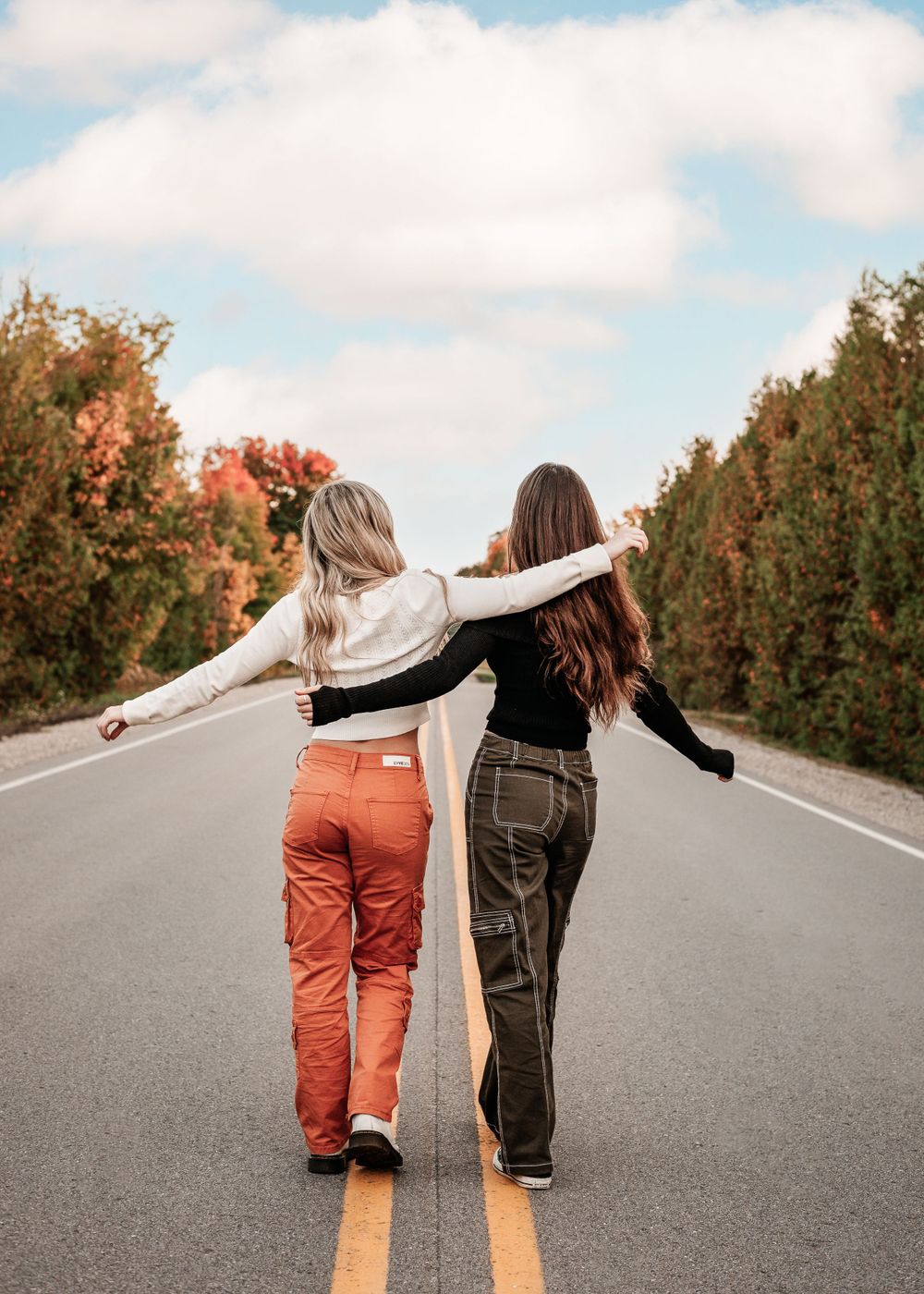 Two friends wearing colorful pants walk together arm in arm down the middle of an empty road with autumn trees along the sides.
