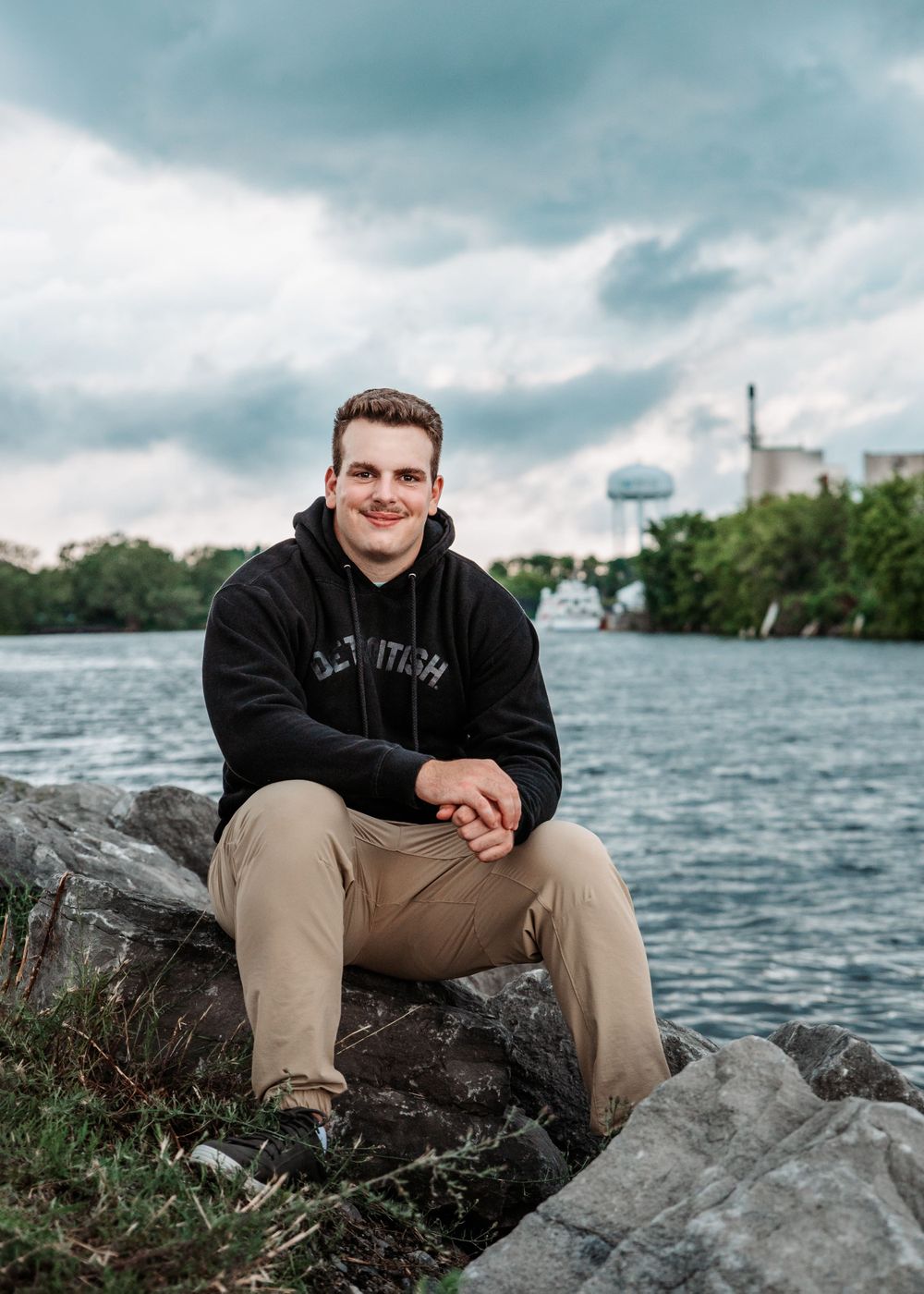 Person in black hoodie sits casually on rocky shoreline with water and industrial buildings visible in background.