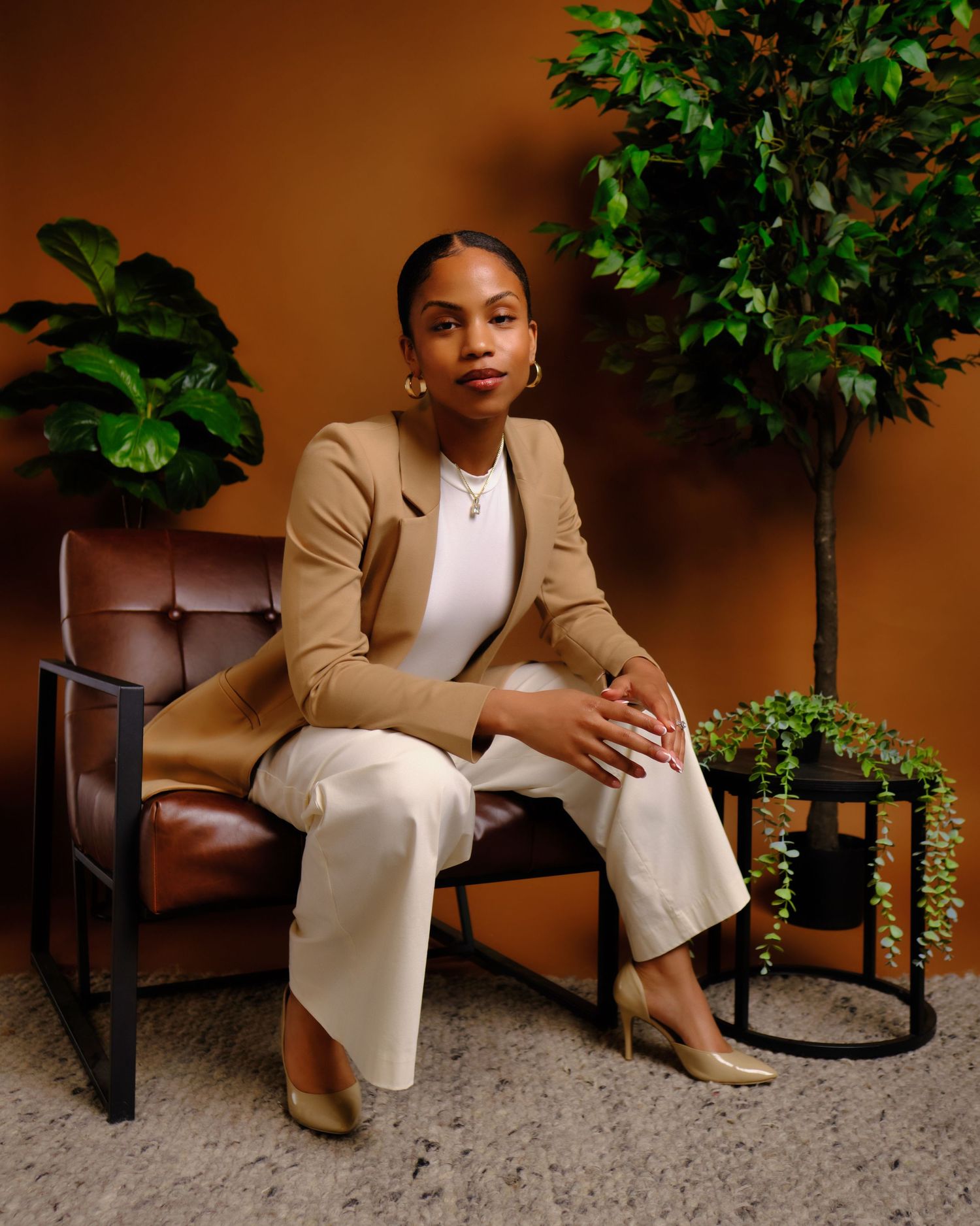 Professional wearing tan blazer and white pants sits elegantly in leather armchair.