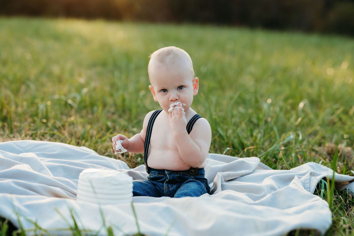 A baby sits on a white blanket in the grass during golden hour outdoor photography session.