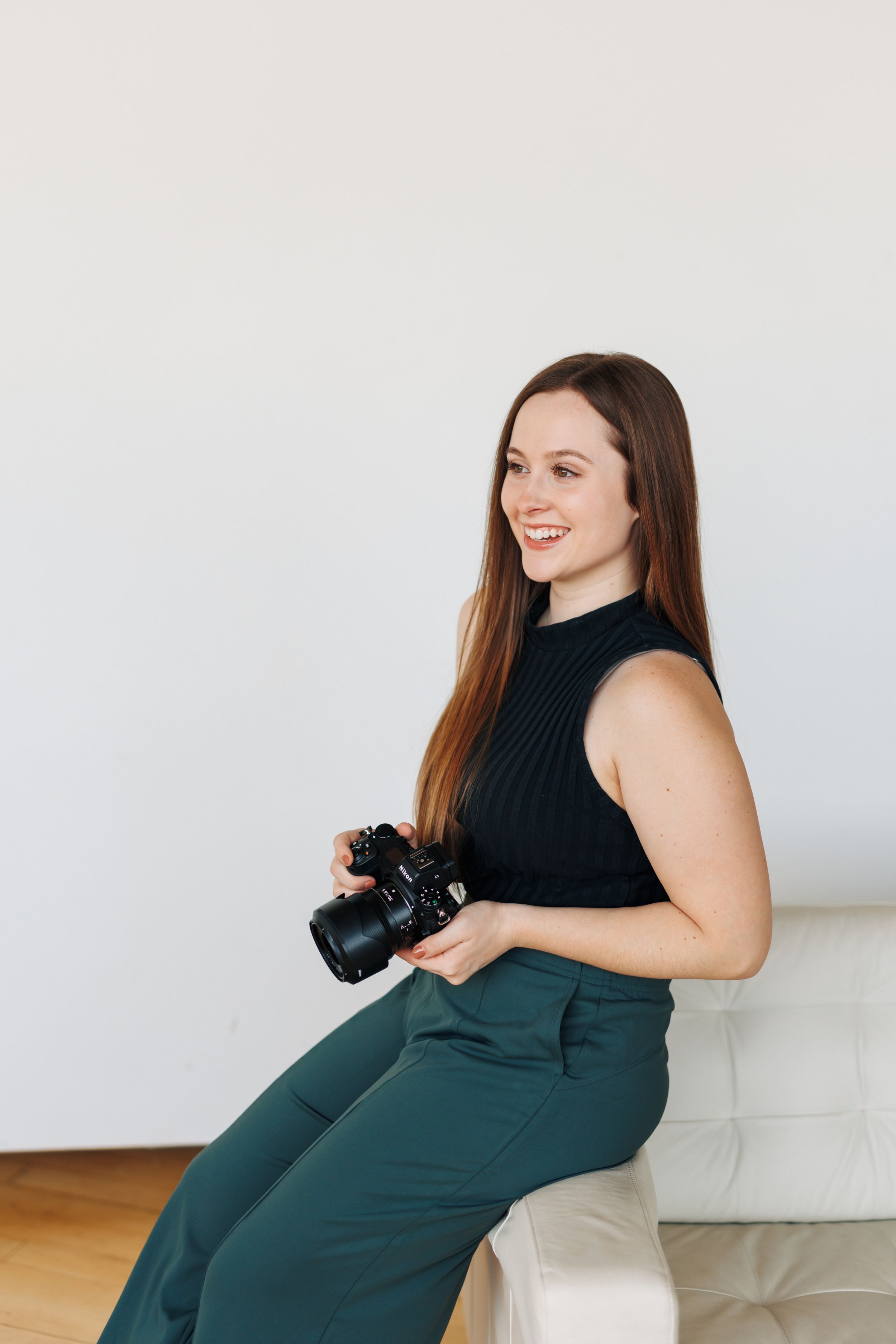 Professional photographer in a black sleeveless top and green pants holds a mirrorless camera while sitting.