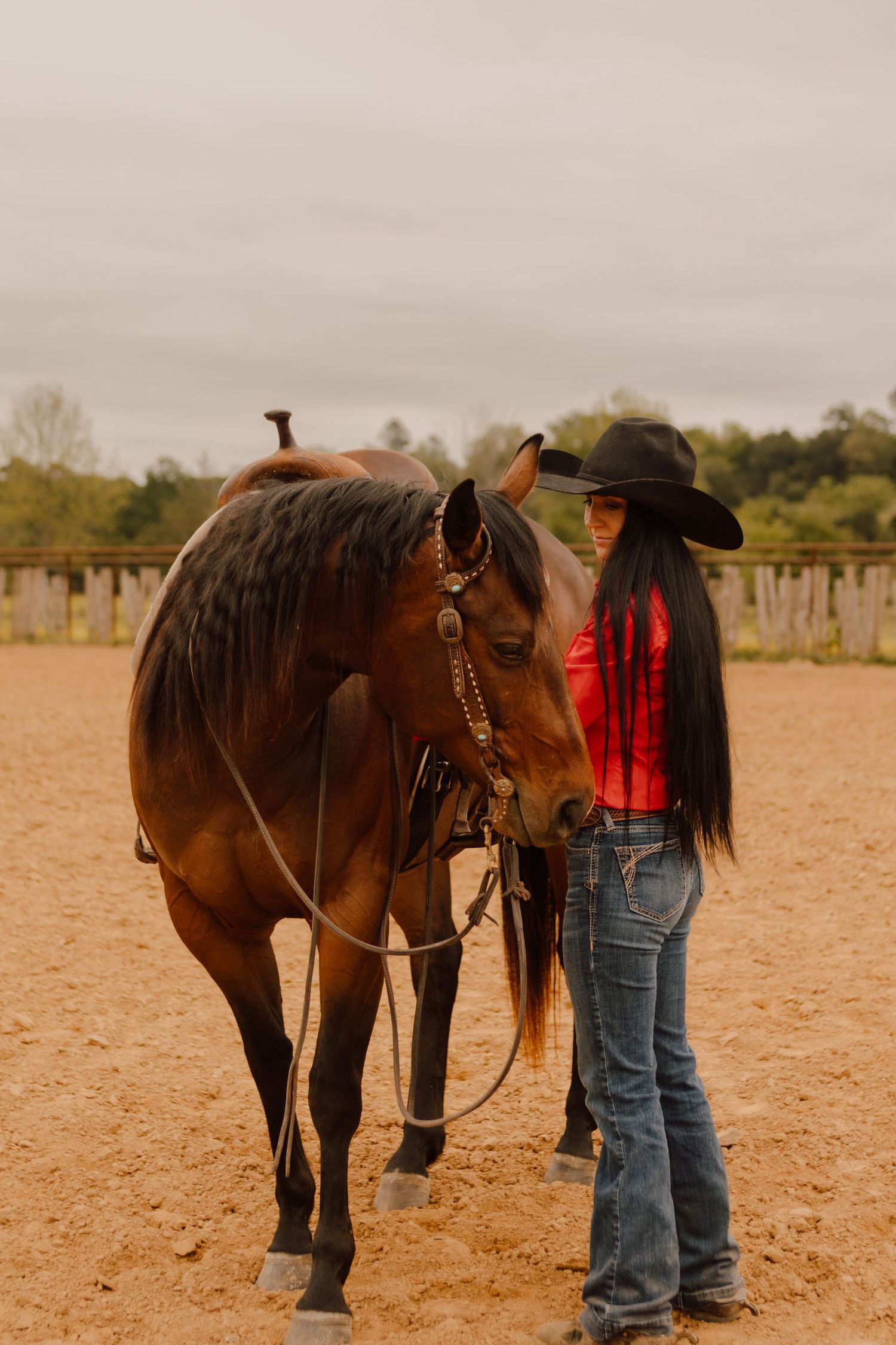 Person in western wear standing with a brown horse in a sandy arena.