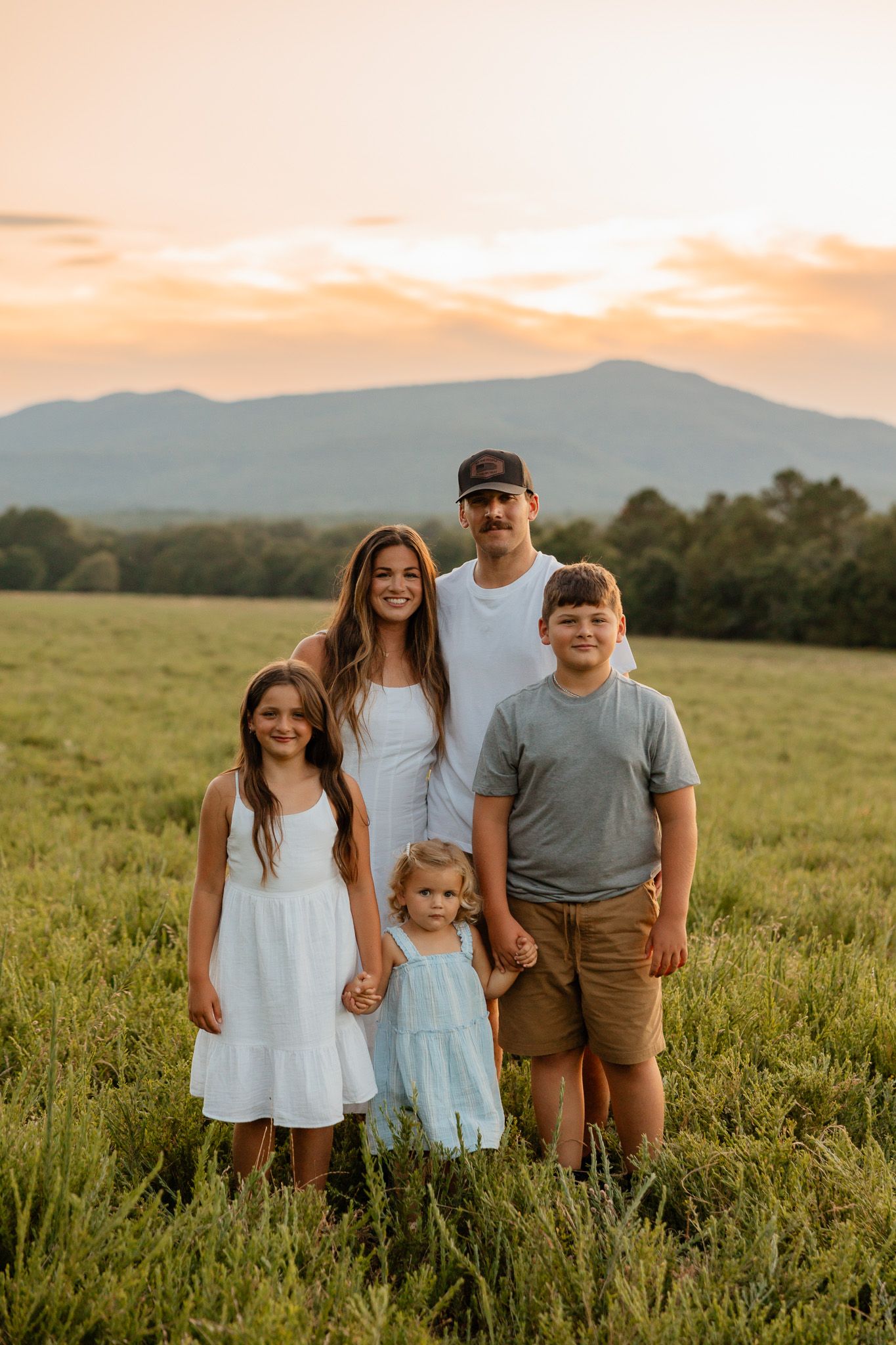 A family stands together in a grassy field at sunset with mountains visible in the background.