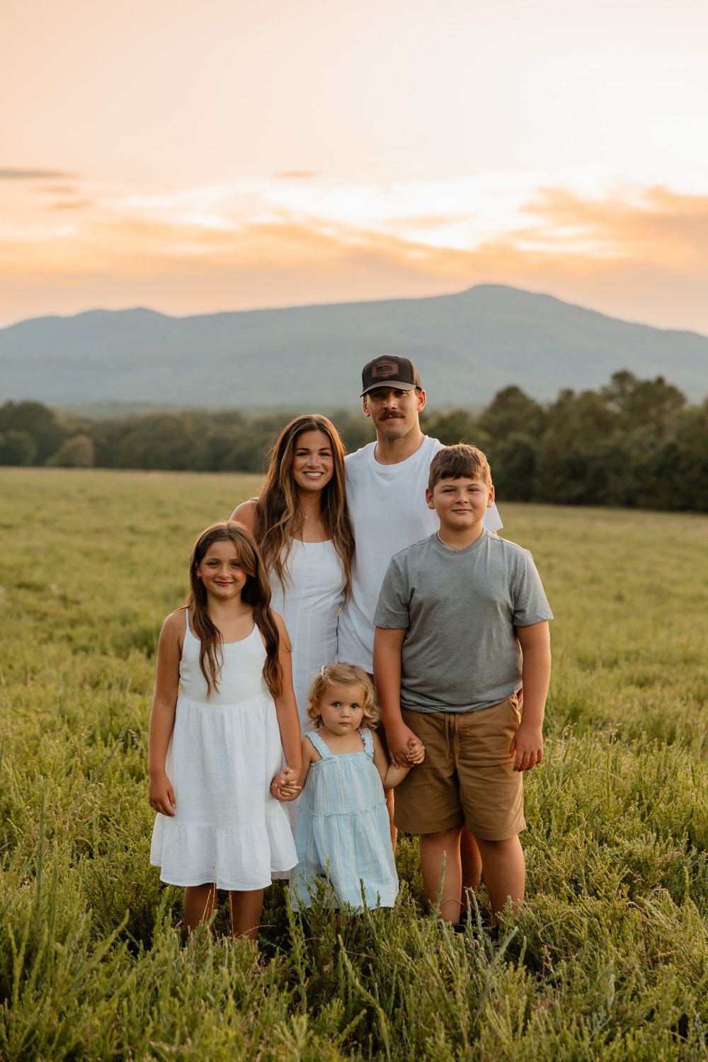A family stands together in a grassy field at sunset with mountains visible in the background.