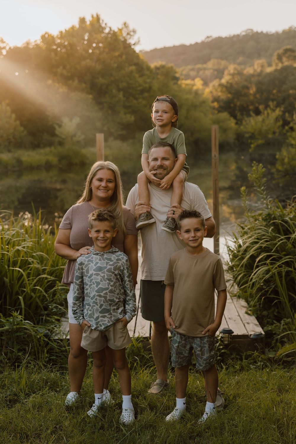 A family poses together at sunset on a grassy path near a pond surrounded by lush summer greenery.