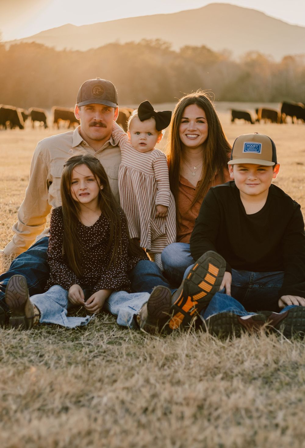 Family sitting together in field at sunset with mountain landscape and grazing cattle.