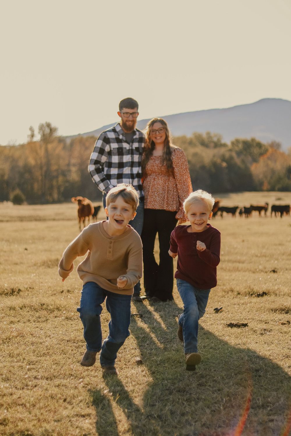 Children running joyfully in field with cattle and mountains in background at sunset.