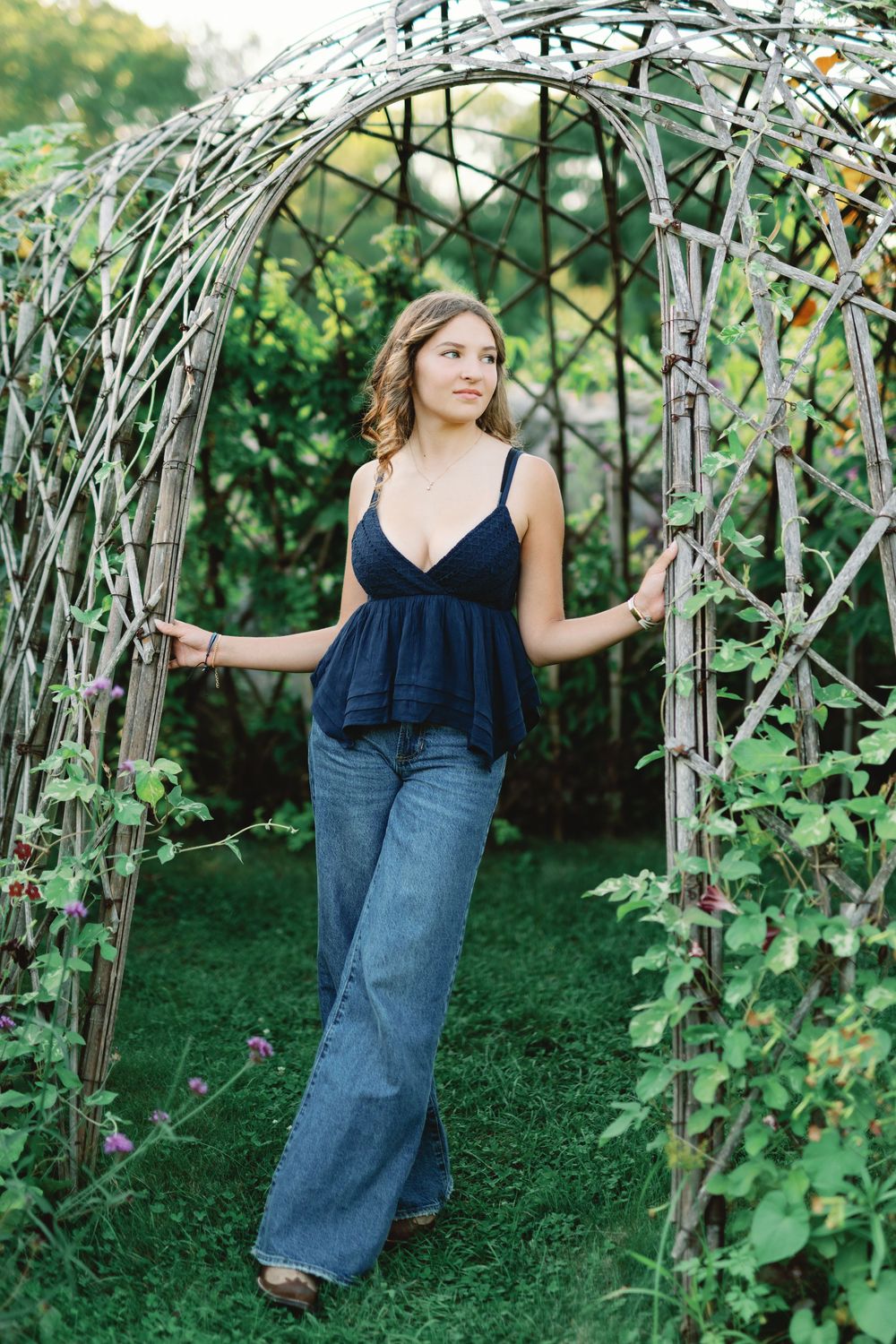 Someone poses by a rustic wooden garden arbor with climbing vines and greenery in a natural outdoor setting.