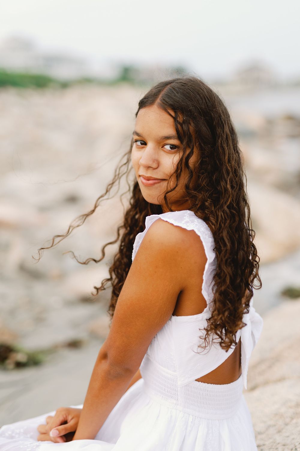 Young child in white sundress poses near beach with windblown curly hair against blurred coastal background.