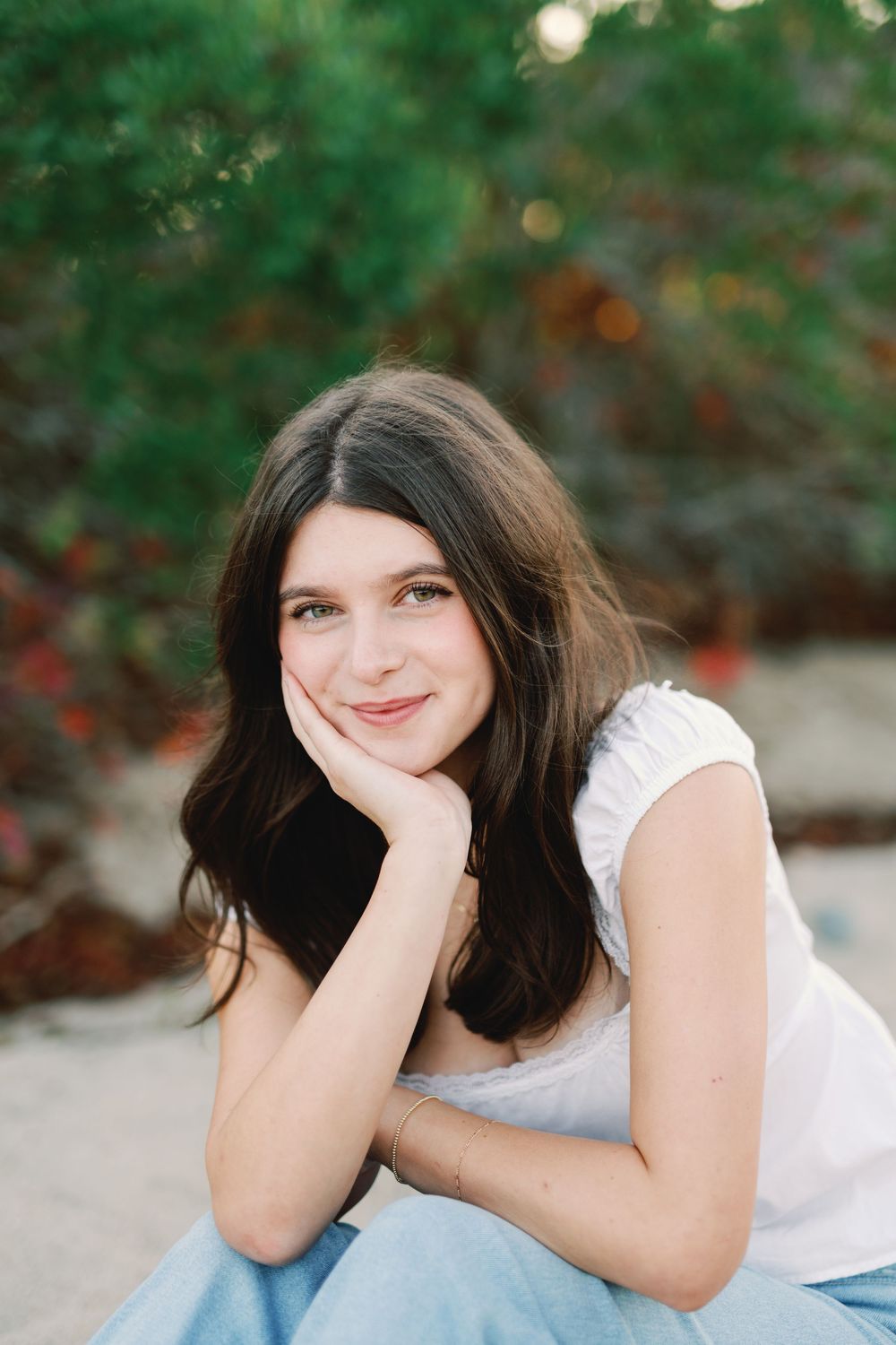 A smiling person in a white sleeveless top poses outdoors with their chin resting on their hand against a green nature backdrop.