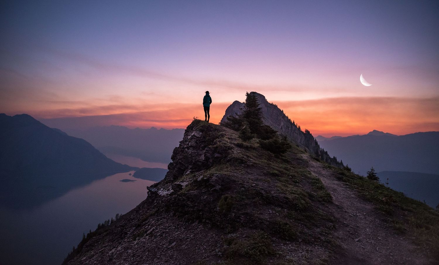 Silhouette of a hiker standing on a mountain ridge against a purple sunset sky with crescent moon.
