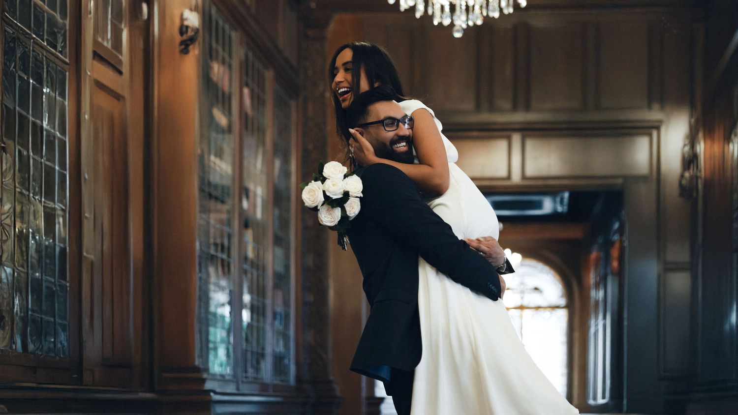 A couple in formal wedding attire embrace in an elegant hallway with wooden paneling and a chandelier overhead.