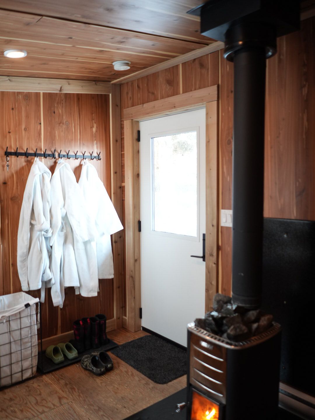 Traditional wood-paneled sauna room with white robes hanging and a black heating stove.