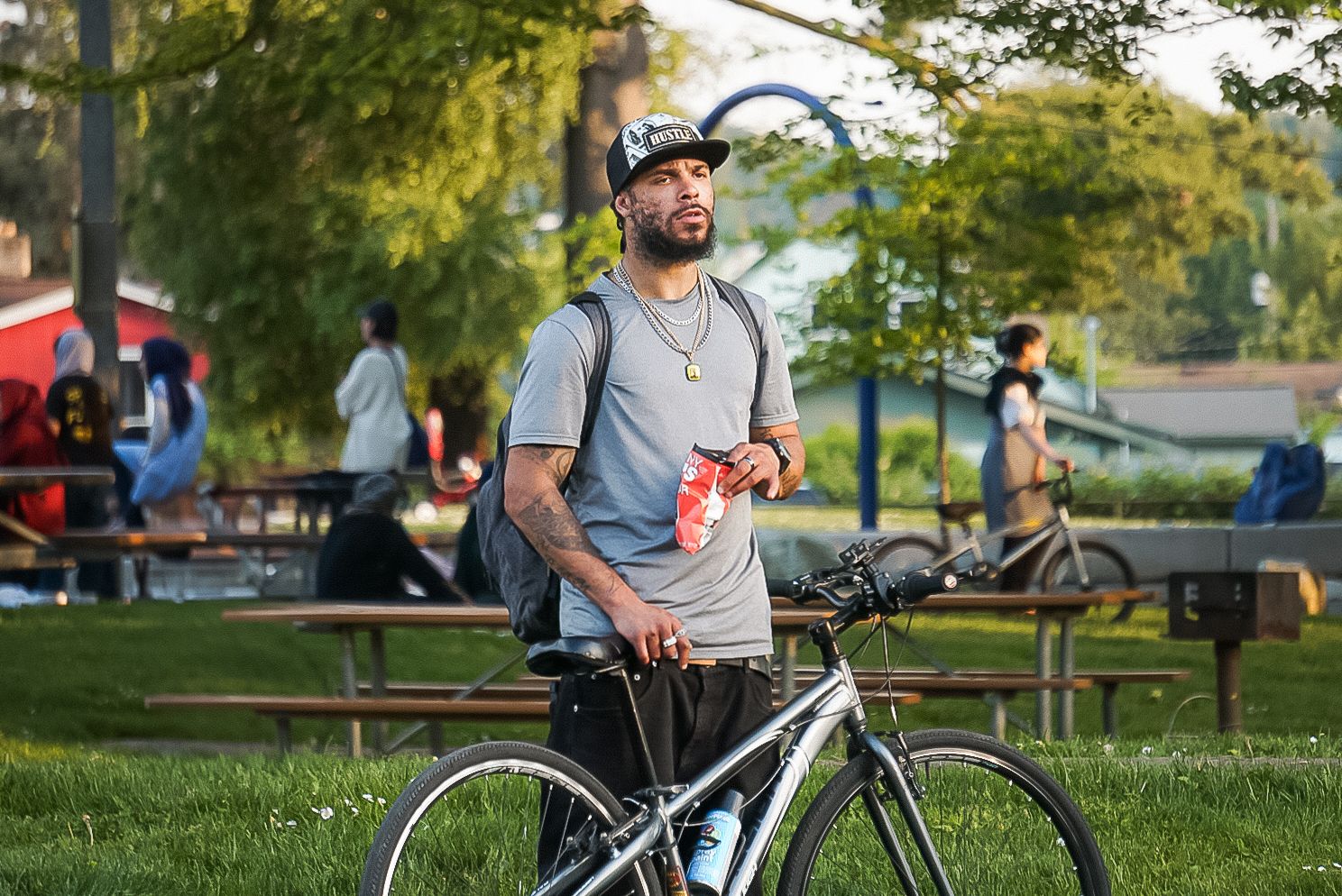 Cyclist in casual attire standing with mountain bike at sunset in public park with picnic tables and trees.