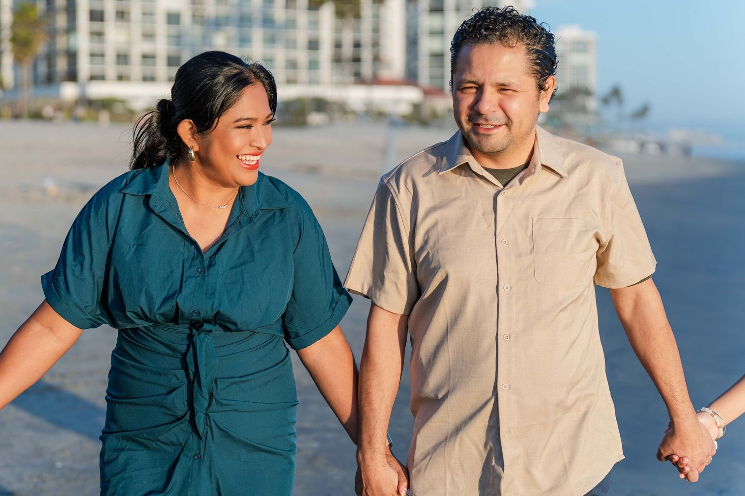 Couple in casual attire smiling on beach with buildings in background.