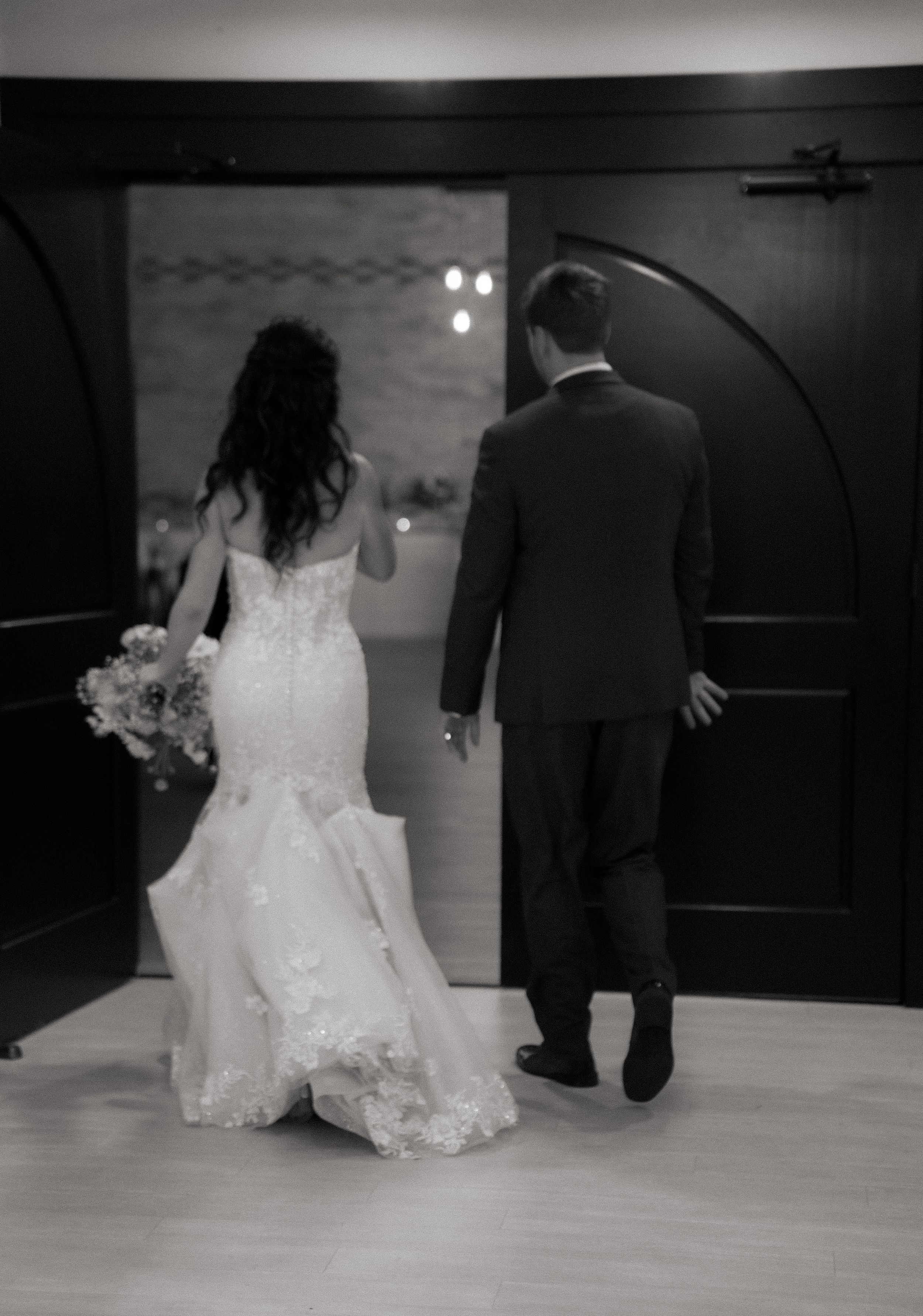Black and white wedding photo showing bride in lace mermaid gown with bouquet and groom in suit walking through doorway.