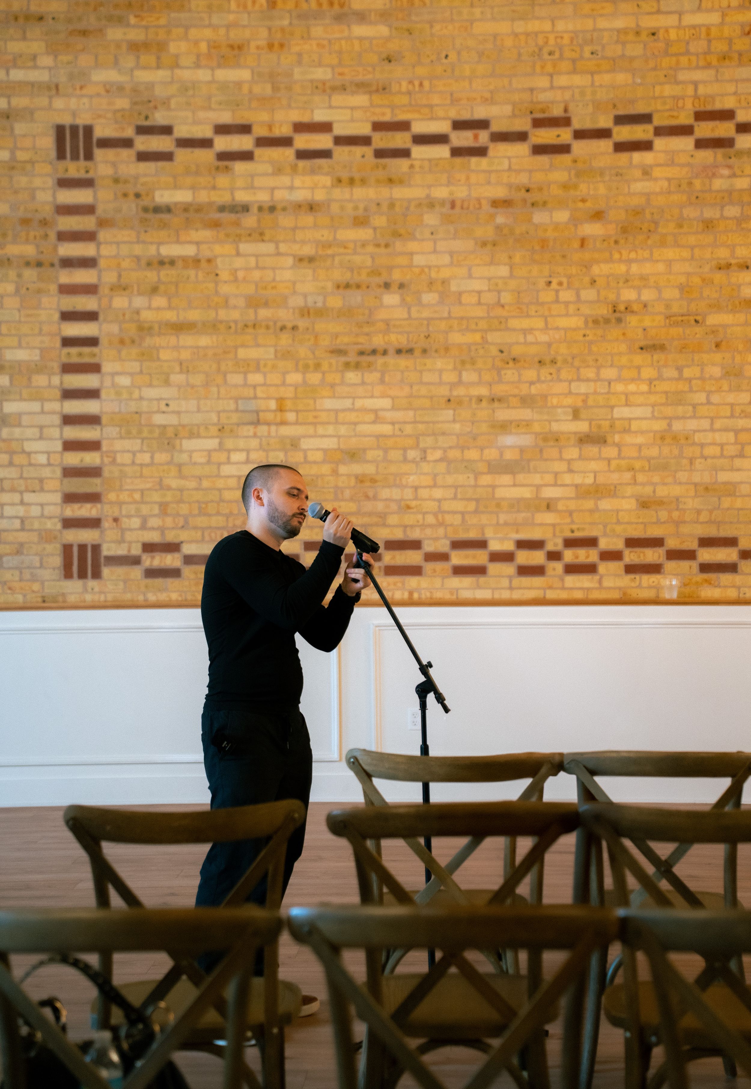 A person in black stands at a microphone in a room with wooden folding chairs and a decorative brick wall pattern.