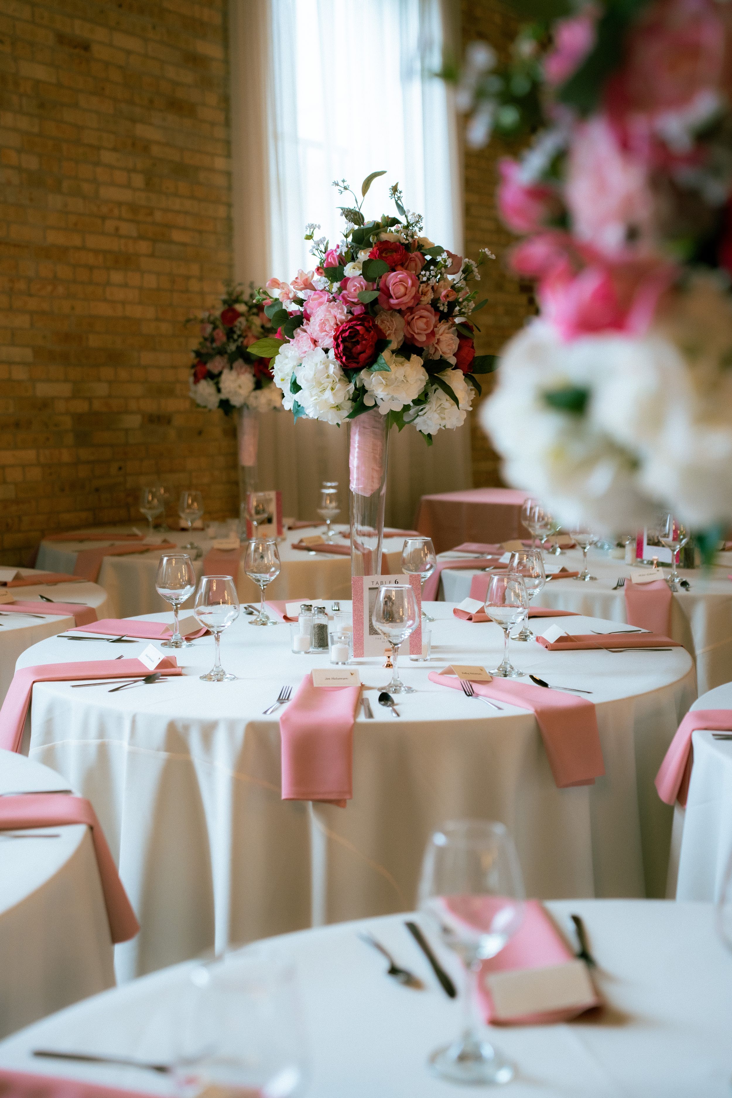 Elegant wedding reception table with pink accents, white linens and tall floral centerpiece against brick wall.