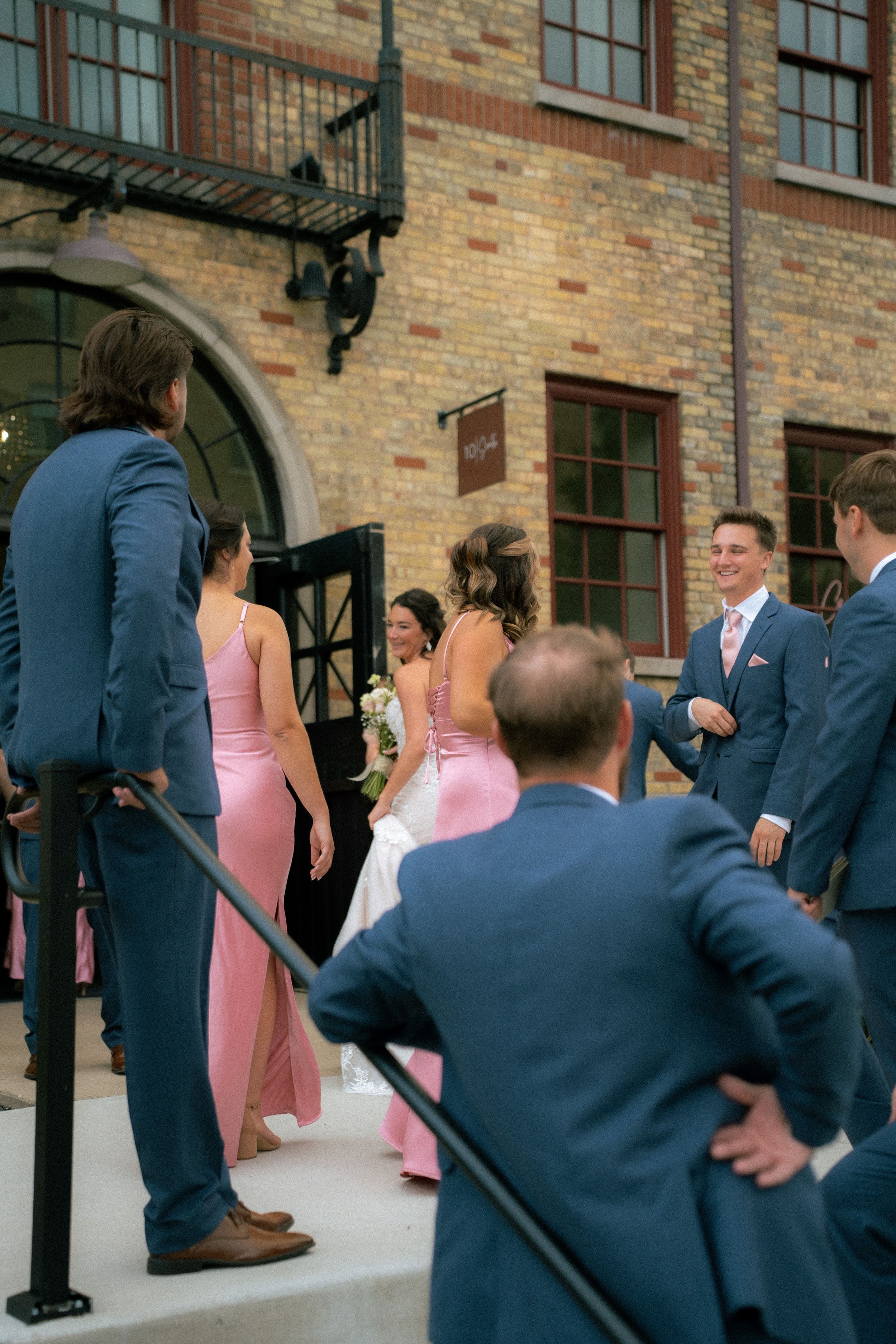 Bridal party in pink dresses and navy suits gathers on steps outside a brick building with arched doorway.