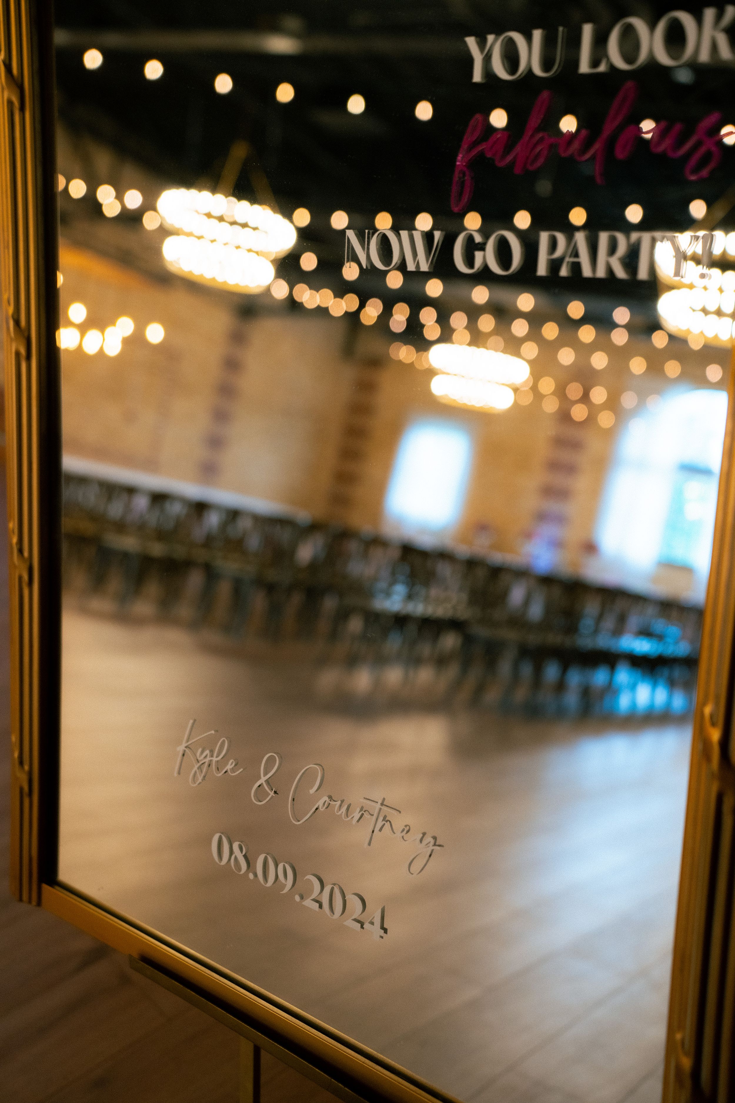 Wedding venue mirror displays 'You look... & company' text with date 08.09.2021 against blurred reception hall with string lights.