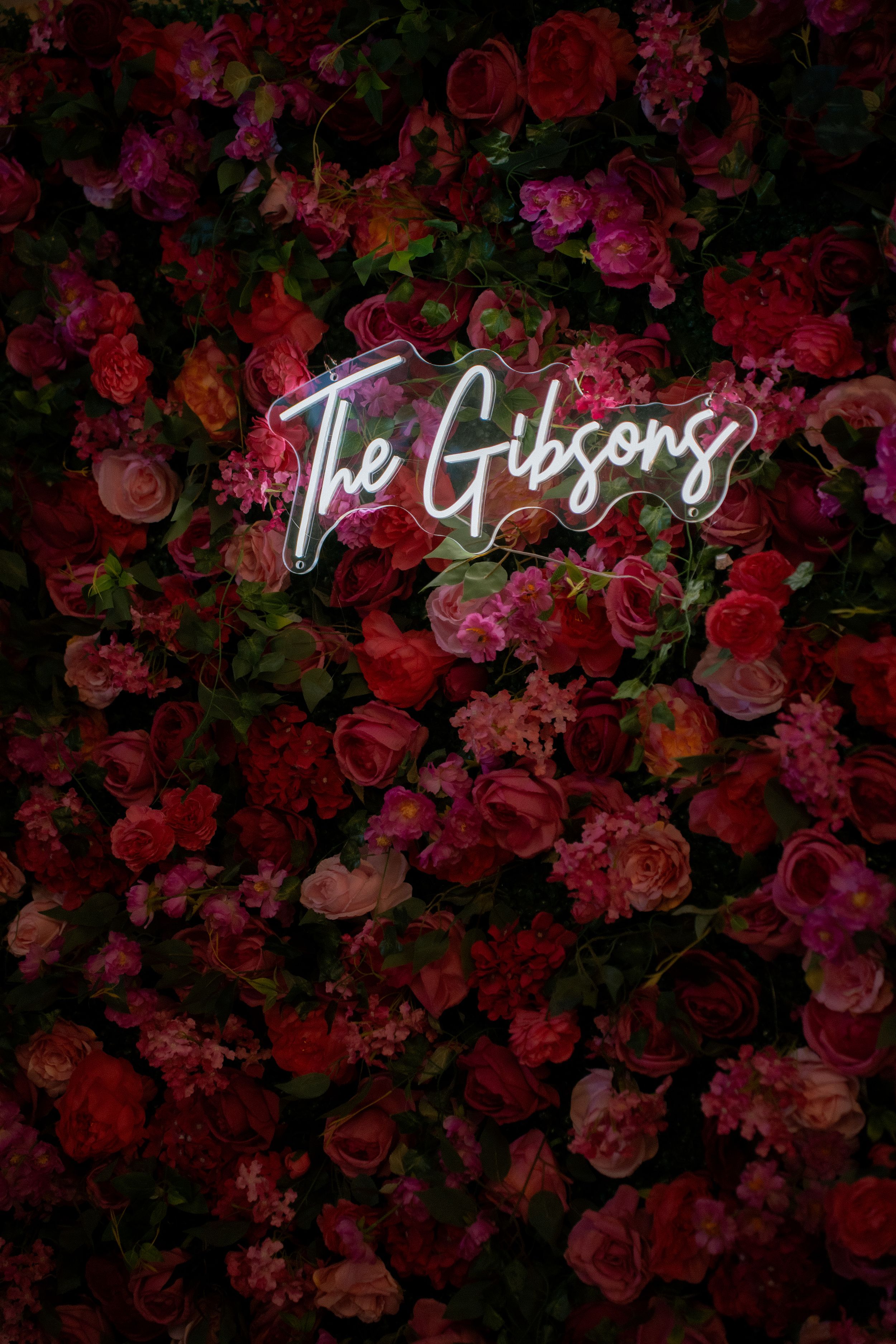 Neon sign reading 'The Gibsons' illuminated against a dark background of red and pink roses and flowers.