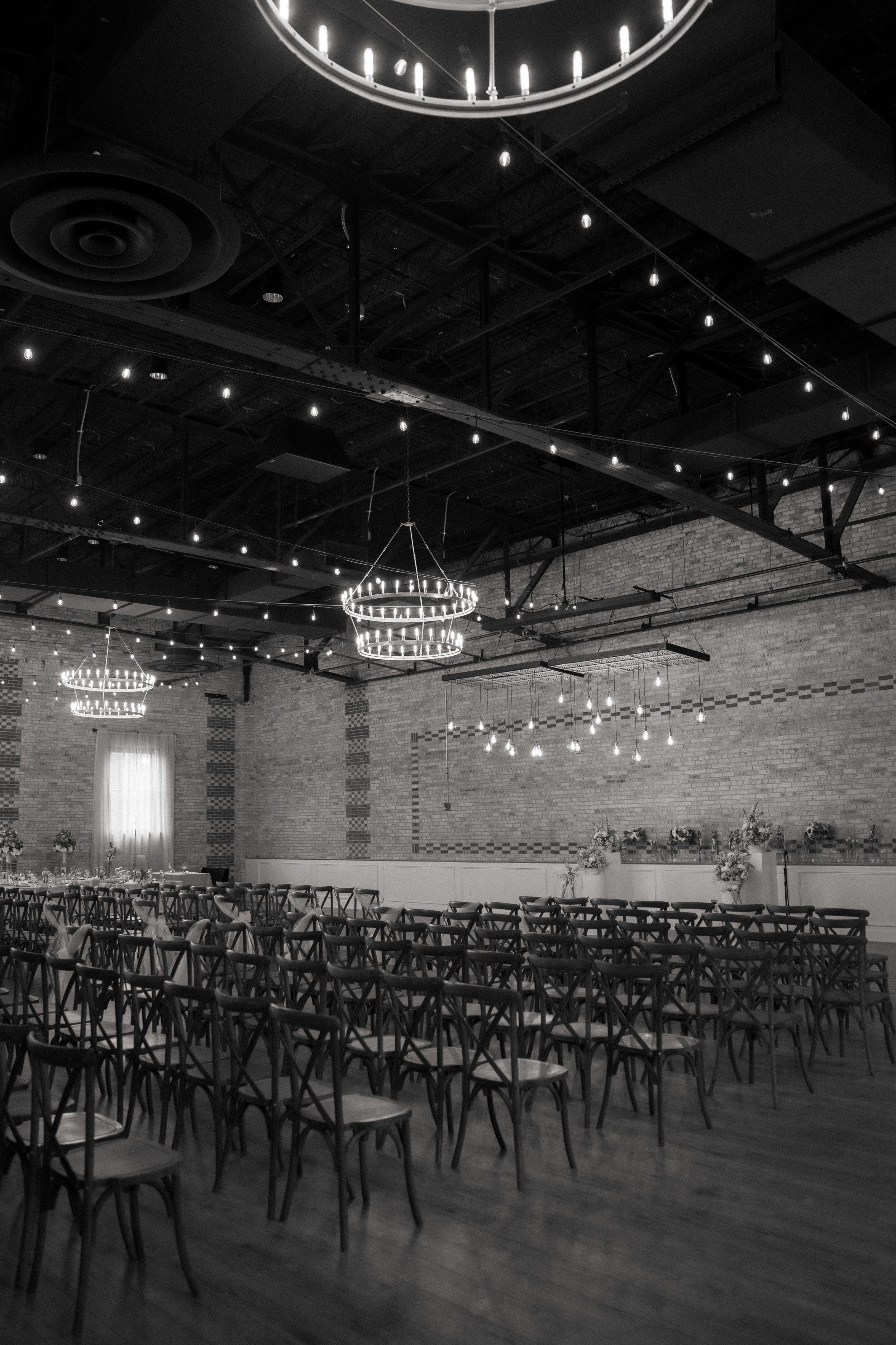 Elegant event venue interior featuring crystal chandeliers, exposed brick walls and rows of cross-back chairs on hardwood floors.