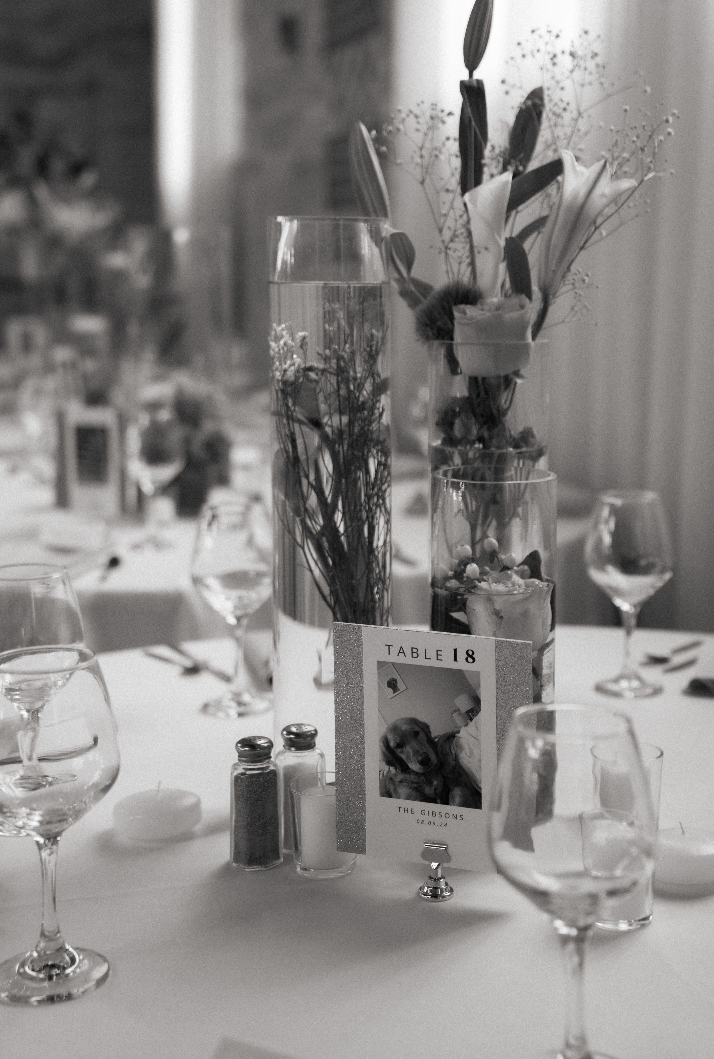 Black and white photograph of an elegant table setting with floral centerpieces in glass vases and wine glasses.