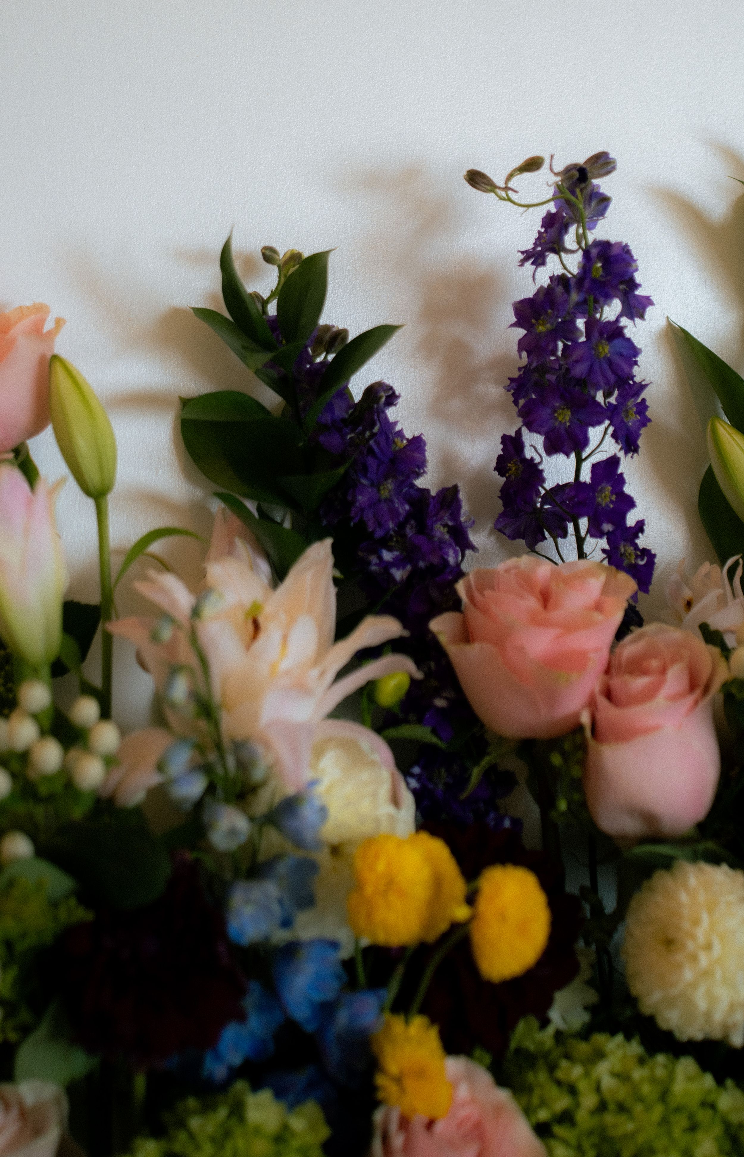 A floral arrangement with pink roses, purple delphinium, yellow billy balls, blue flowers and green foliage against a light background.
