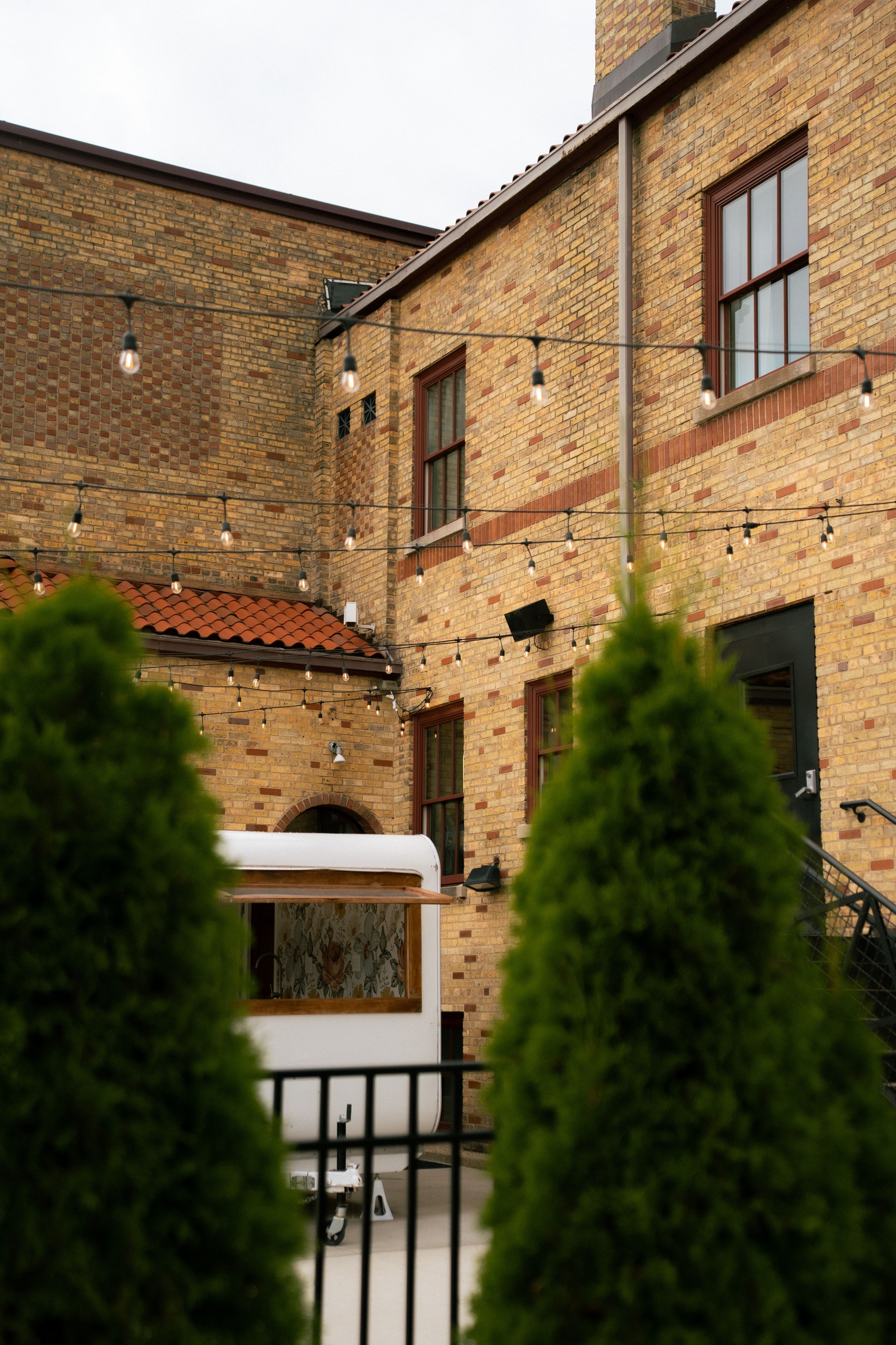 Exterior view of brick building with windows and evergreen shrubs in foreground behind metal railing.
