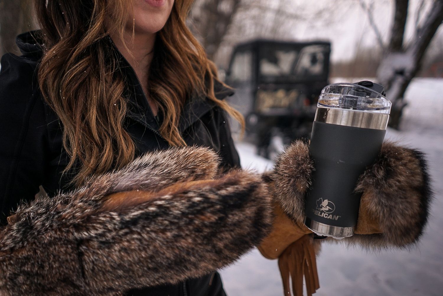 Person holding insulated tumbler while wearing fur mittens in winter setting.