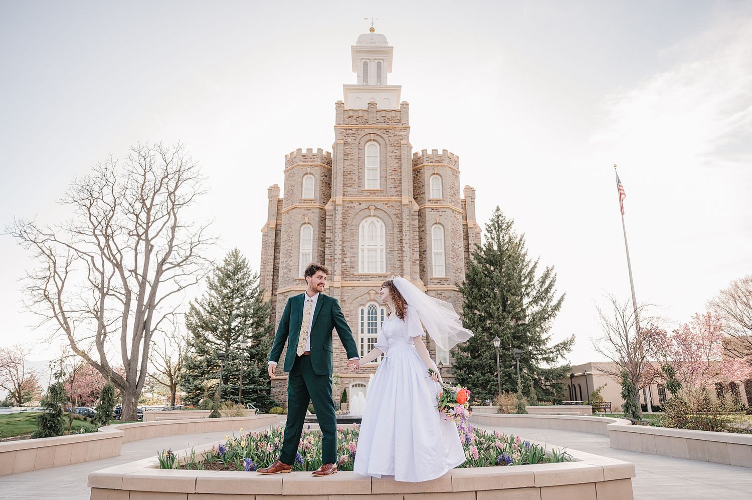 A couple in wedding attire stand together on a pathway in front of a grand Gothic-style Mormon temple on their wedding day.