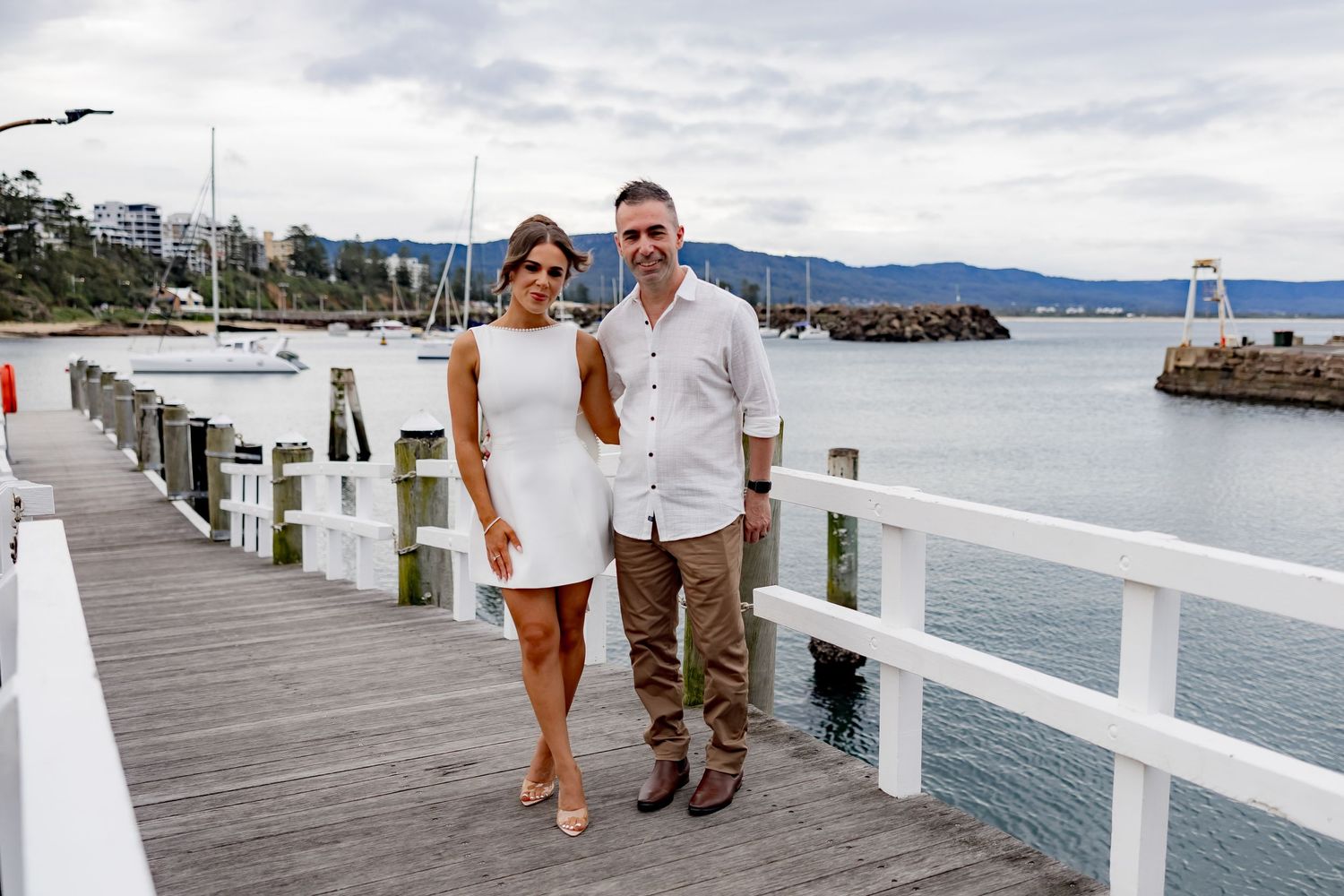 A couple poses together on a wooden pier overlooking a marina with mountains in the distance.