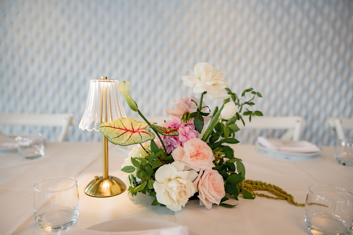 Elegant table centerpiece featuring a small gold lamp and pink and white roses with greenery on white tablecloth.