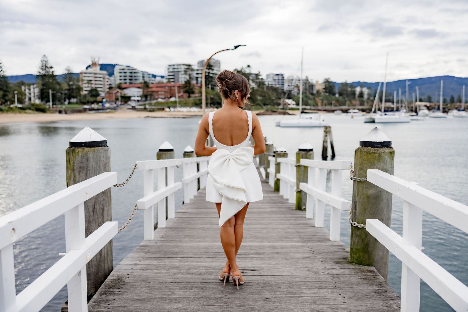 A white-dressed person walks on a wooden pier overlooking a marina with boats and mountains in the background.