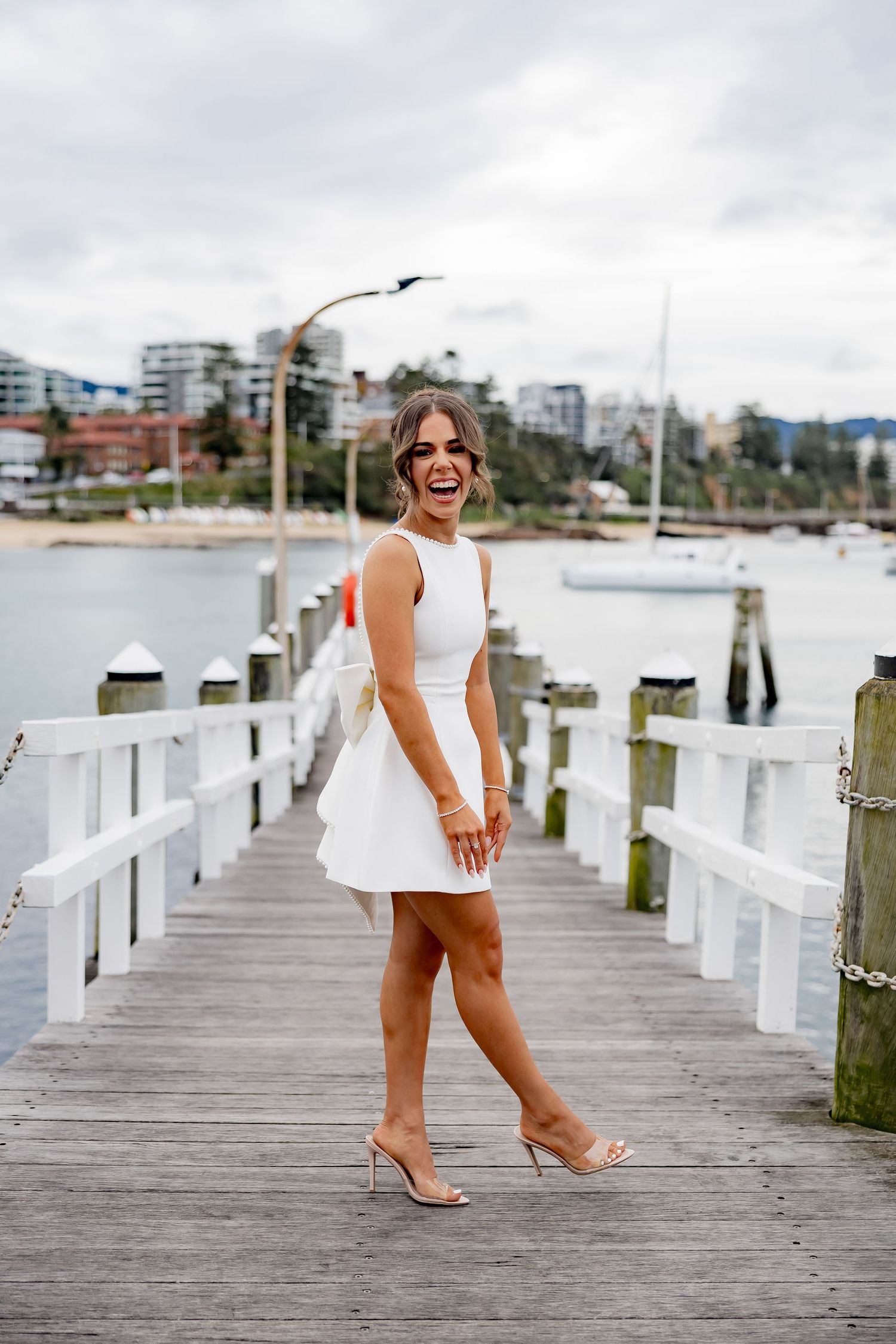 A stylish woman in a white mini dress poses joyfully on a wooden pier by a marina.