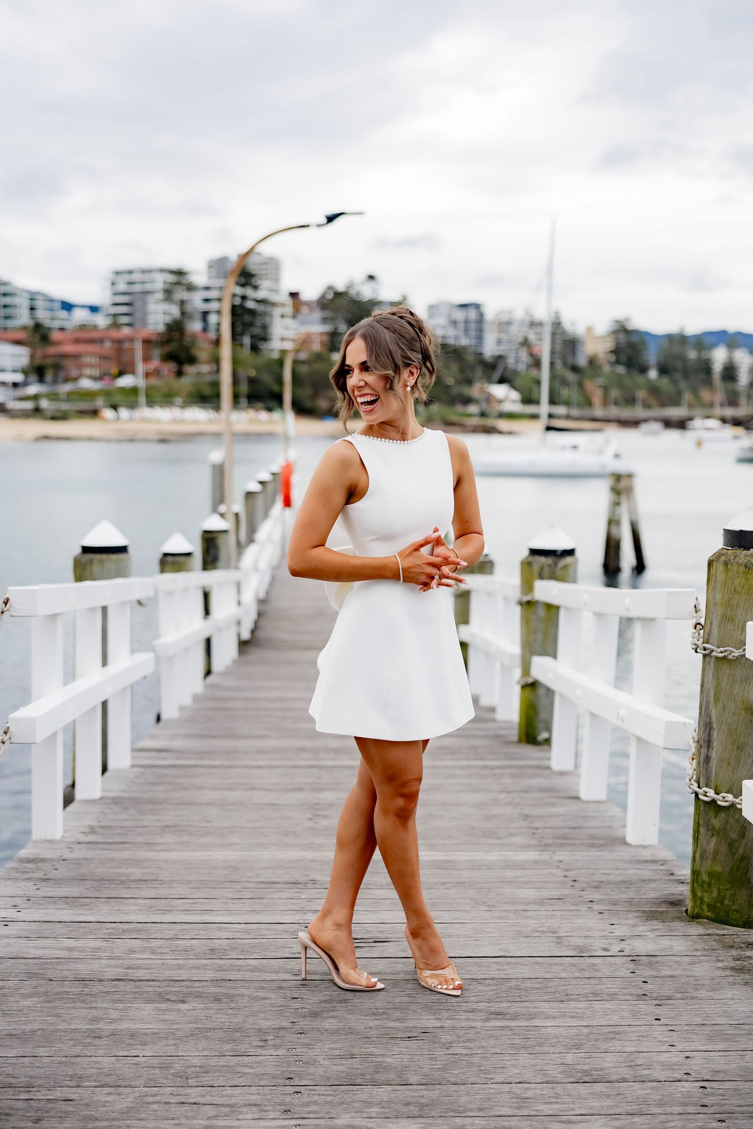 A person in a sleek white summer dress and heels poses confidently on a wooden marina dock with boats in the background.