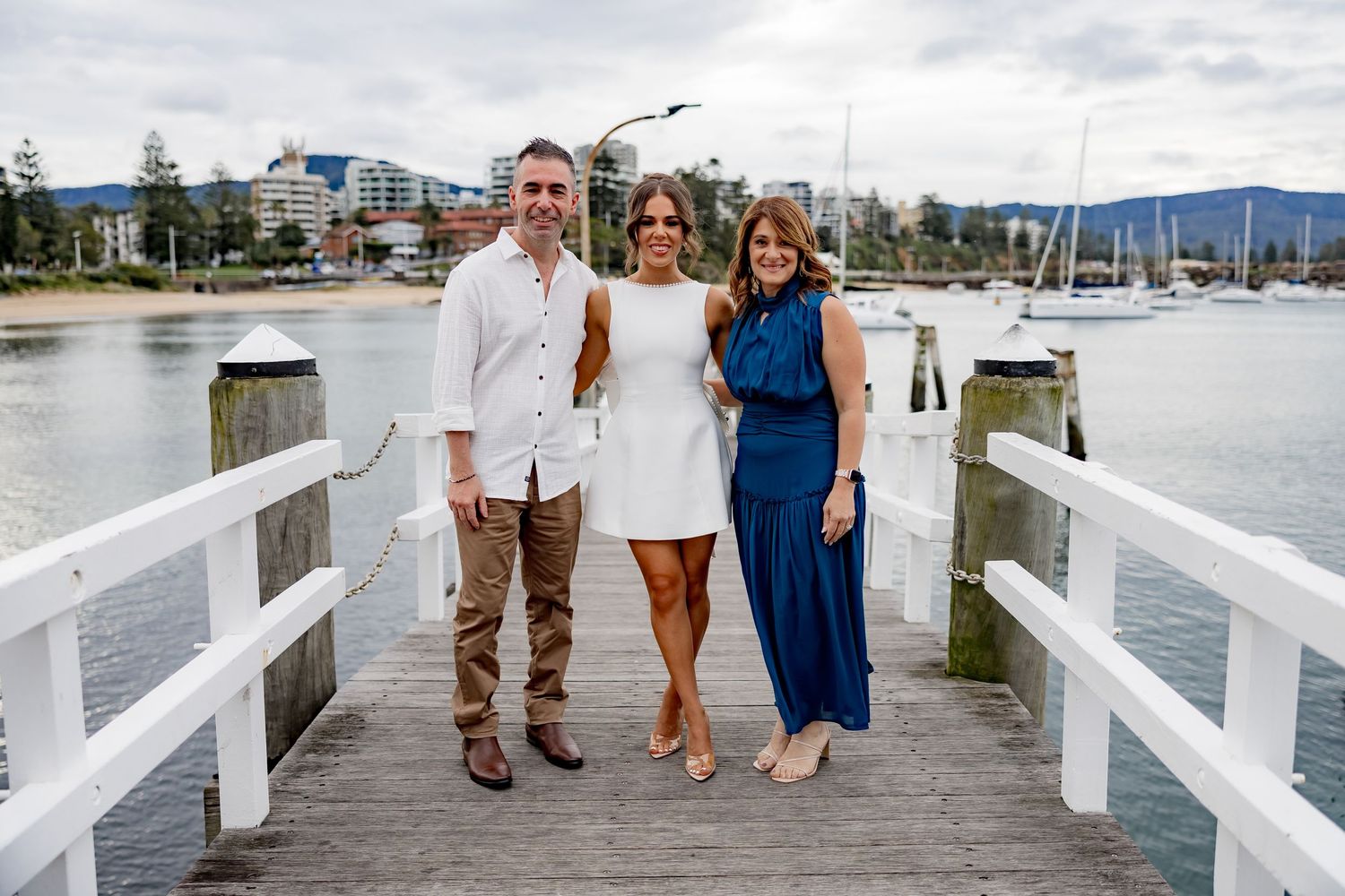 Three people pose together on a white wooden pier with boats and mountains visible in the marina background.