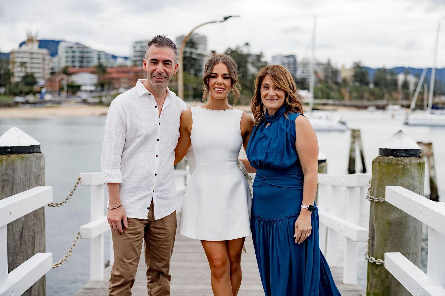 Three people stand together on a waterfront dock with boats and buildings visible in the background on a sunny day.