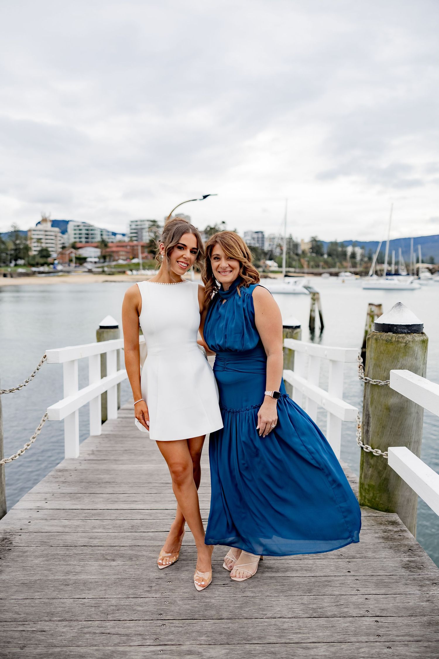 Two women in white and blue dresses pose together on a wooden pier with boats and buildings in the background.