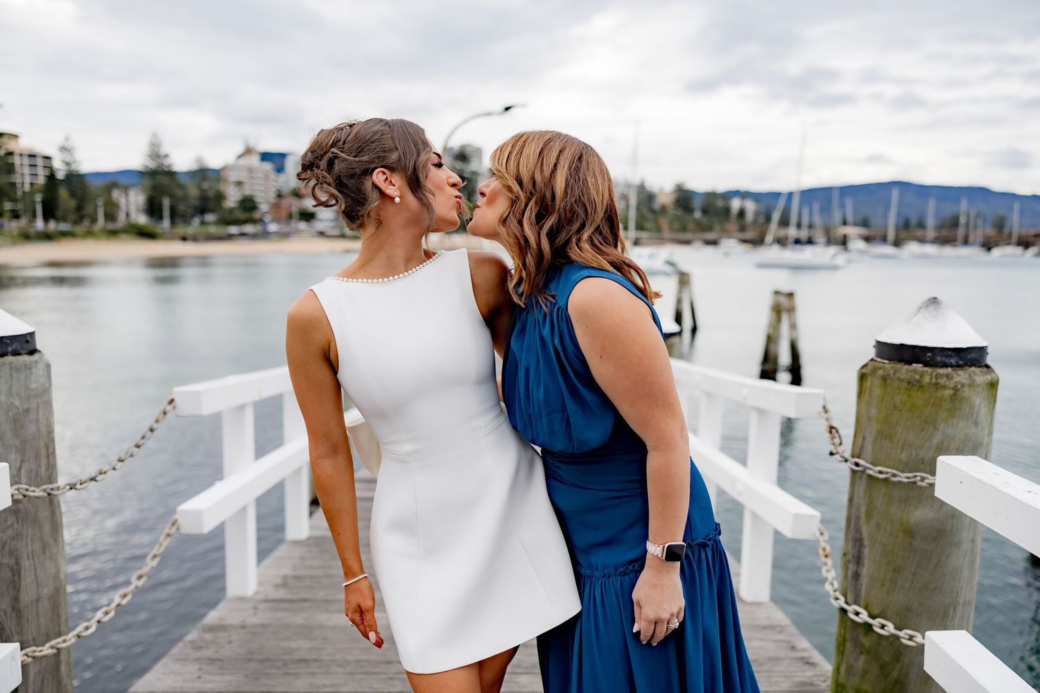 Two people in white and navy dresses share a tender moment on a waterfront dock with mountains in the background.