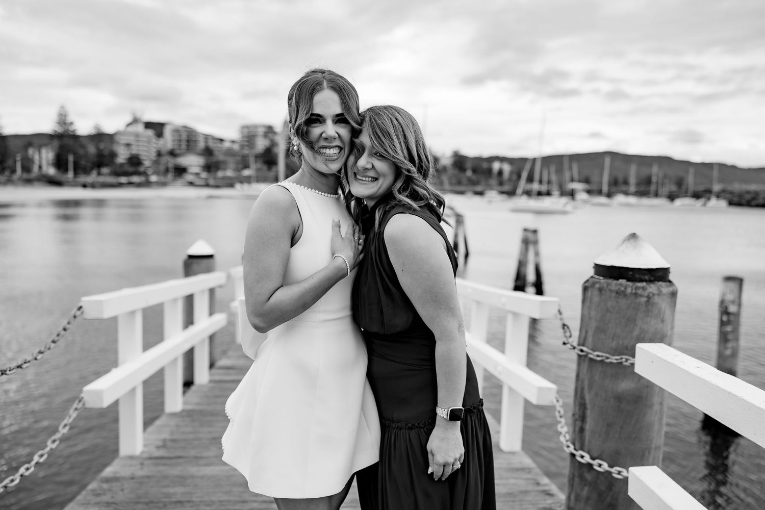 Two people share a joyful moment together on a wooden dock with boats and water in the background.