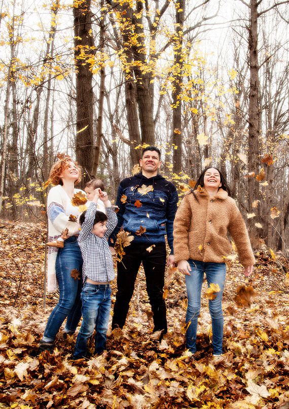 A family plays together in fallen autumn leaves as yellow foliage falls around them in a wooded setting. (Rochester New York)