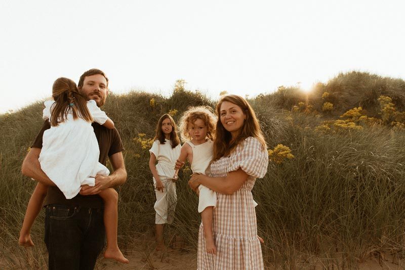 A family poses together in a golden sunlit field during sunset, wearing light colored casual outfits.