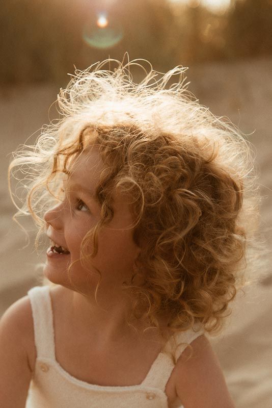 Golden sunlit image of child with wild curly blonde hair wearing white overalls, smiling and looking up at sunset.