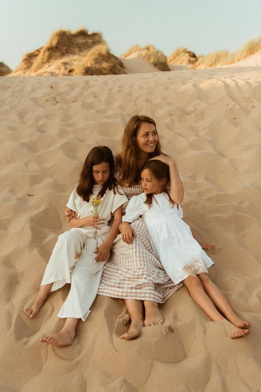 Three people in light-colored dresses and bare feet relax together on sandy dunes at golden hour.
