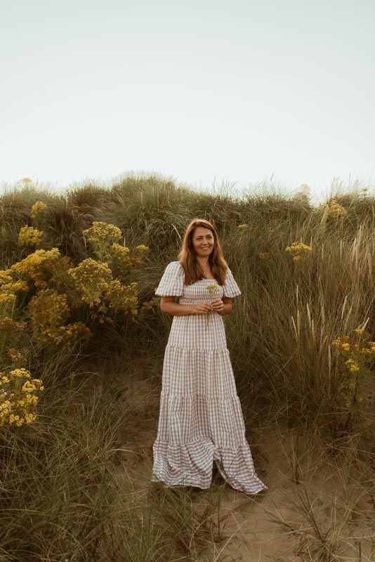 A woman in a white gingham dress stands among tall grass and yellow wildflowers during golden hour.