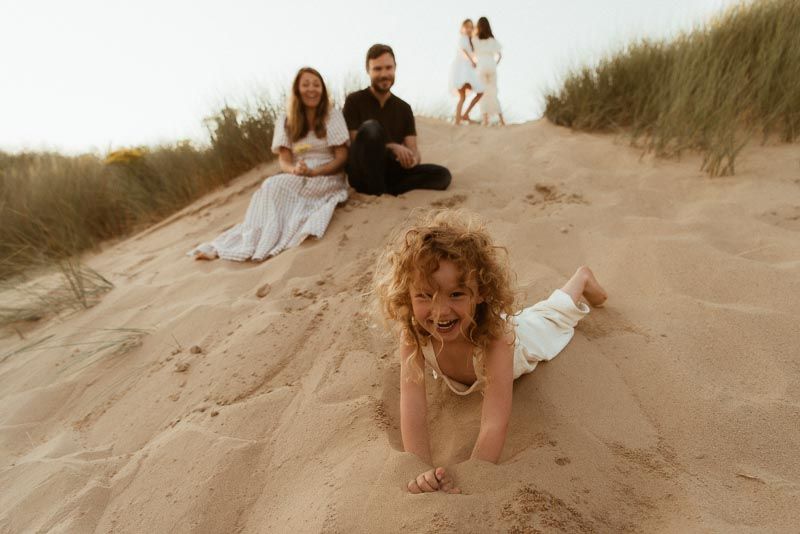 A family sitting on sand dunes at sunset with a playful child crawling in the foreground.