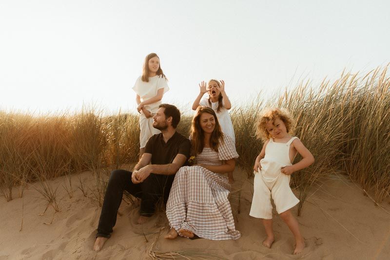 A family sits together on sand dunes during golden hour at the beach with tall grass swaying in the background.