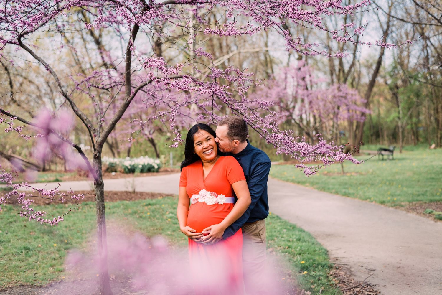 A couple shares an intimate moment under blooming cherry blossom trees in a spring park setting.