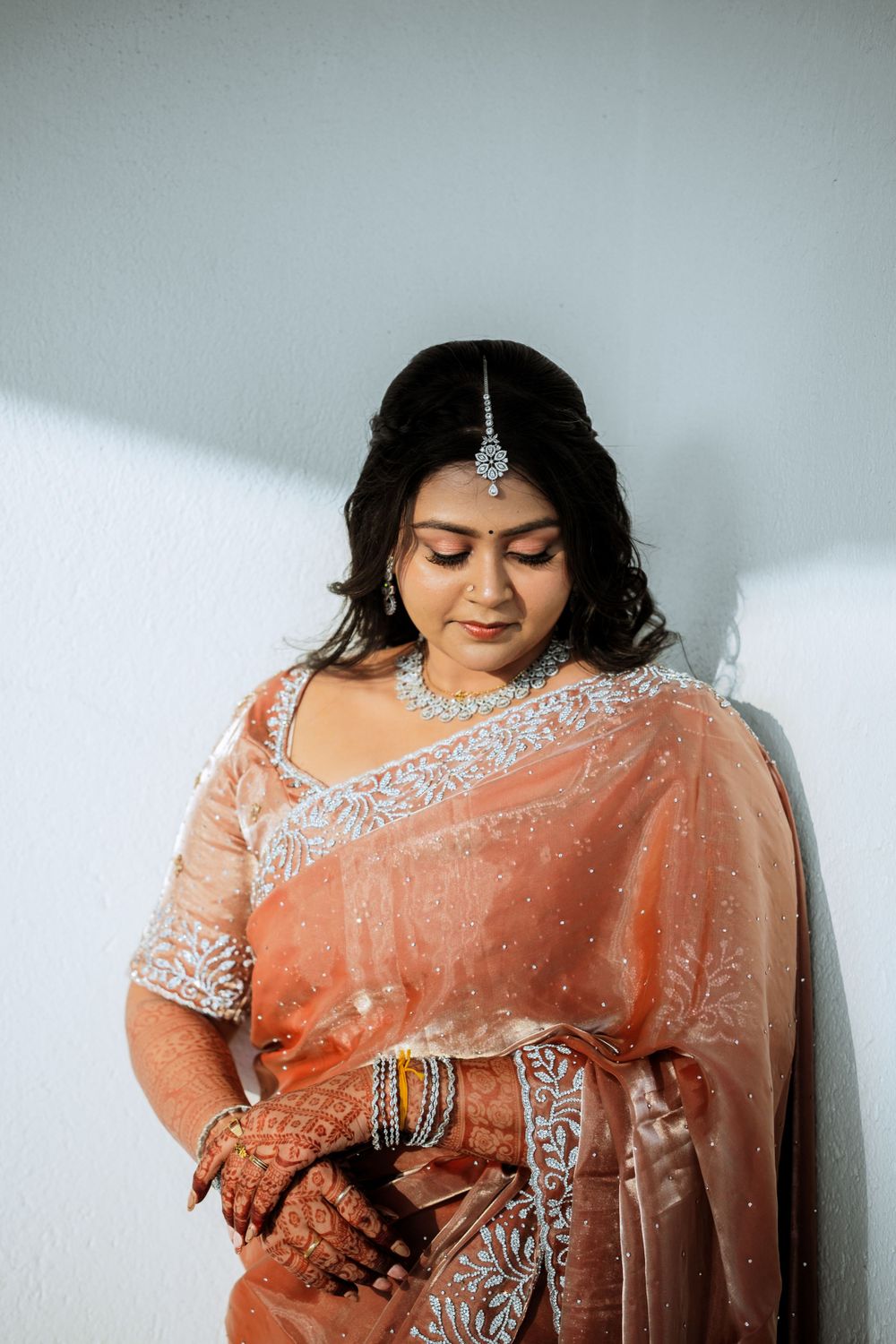 Bride in peach saree with intricate silver embroidery and traditional tikka headpiece looks down gracefully.